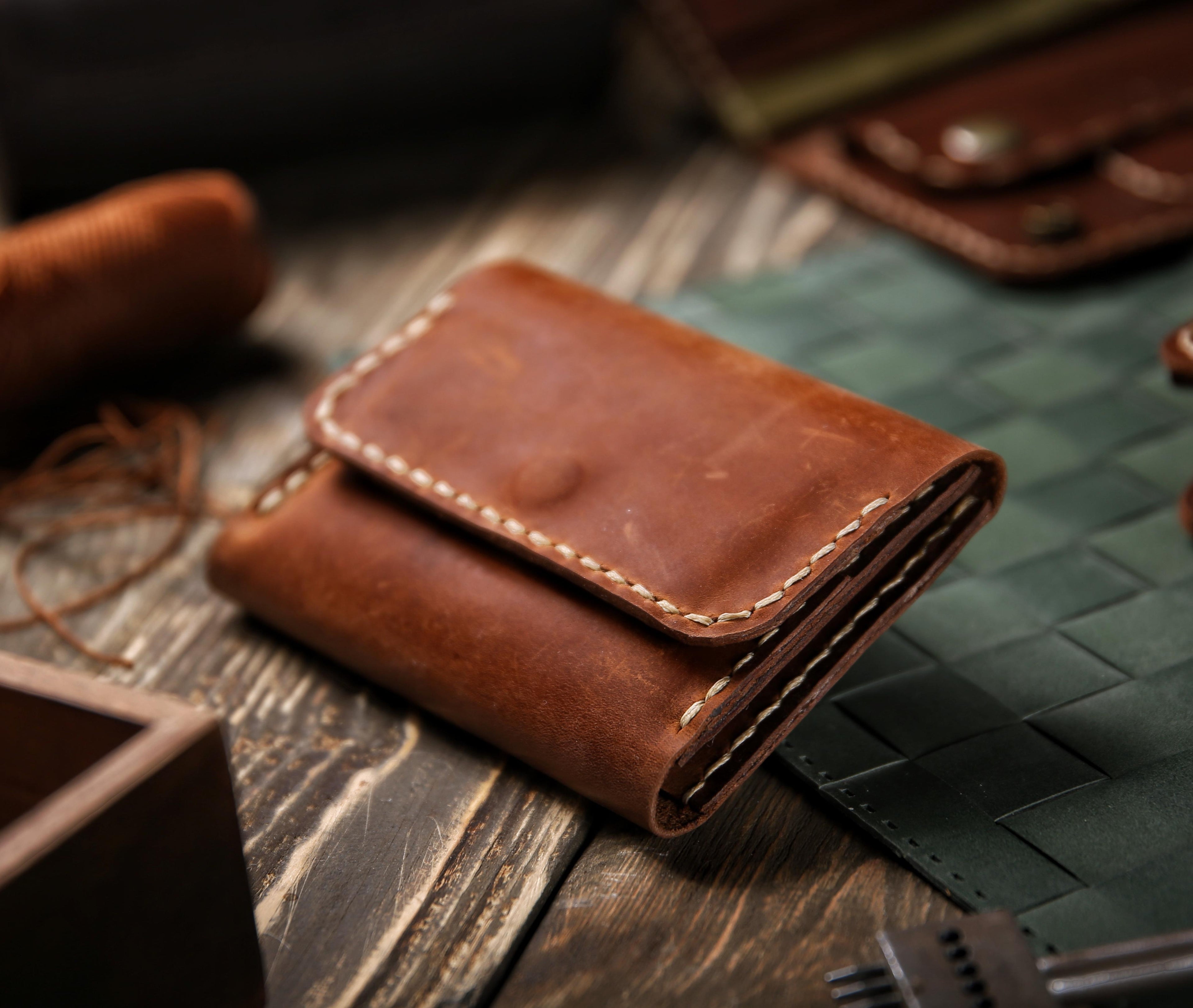 Angled view of a folded compact leather wallet by LIONELL, handcrafted from brown natural leather with prominent white stitching, placed on a wooden table next to leather crafting tools to highlight its vintage character and high quality.