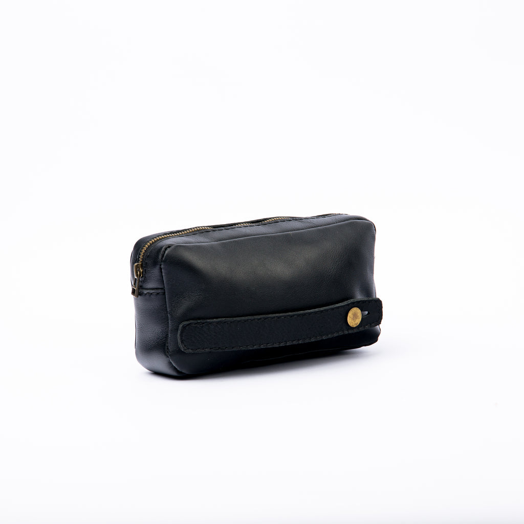 Back view of a LIONELL 'Noir' black leather handbag, showing a built-in leather back strap secured with a brass button for easy hand carrying, featuring a durable vintage design on a white background.