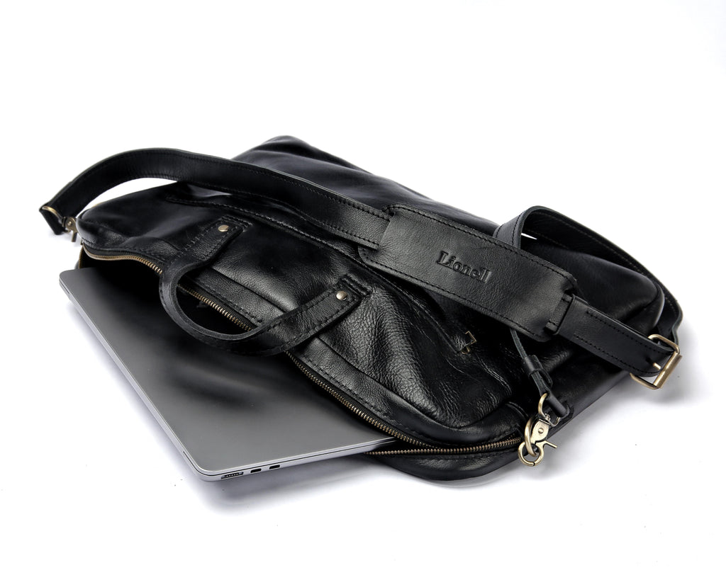 A photo of 'The Noir' pebbled leather laptop bag by LIONELL. Handcrafted from premium black textured leather, showing a laptop partially inside. Features top handles, a padded shoulder strap with the brand logo, and brass hardware, displayed on a white background.