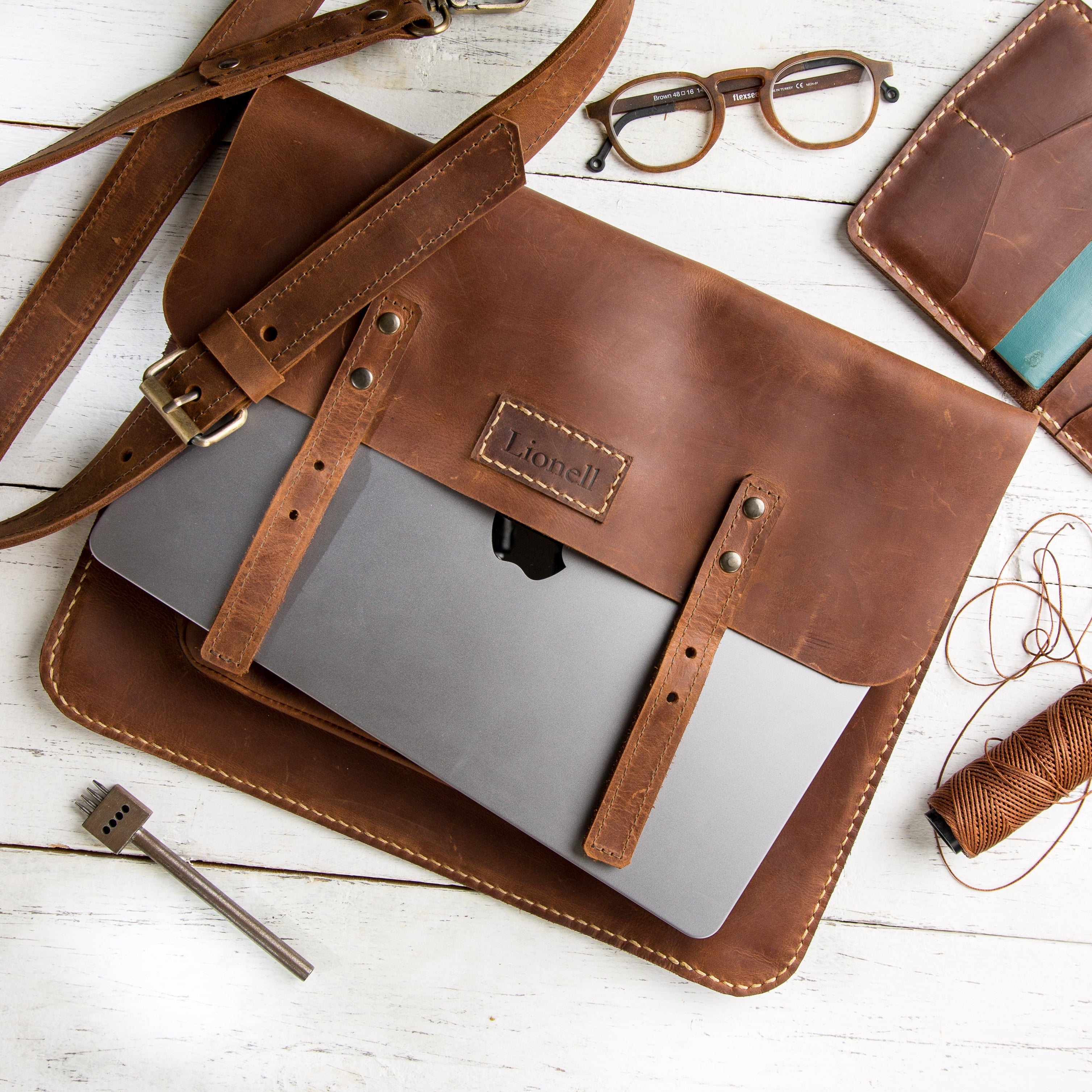 Flat lay of Lionell Rustic Slim Laptop Case in brown leather with laptop, glasses, and tools on white wood.