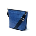 A side-view photo of 'The Urban Crossbag' by LIONELL; handcrafted from rich blue genuine leather, featuring a modern slim design with visible stitching and an adjustable dark blue fabric shoulder strap, displayed on a clean white background.