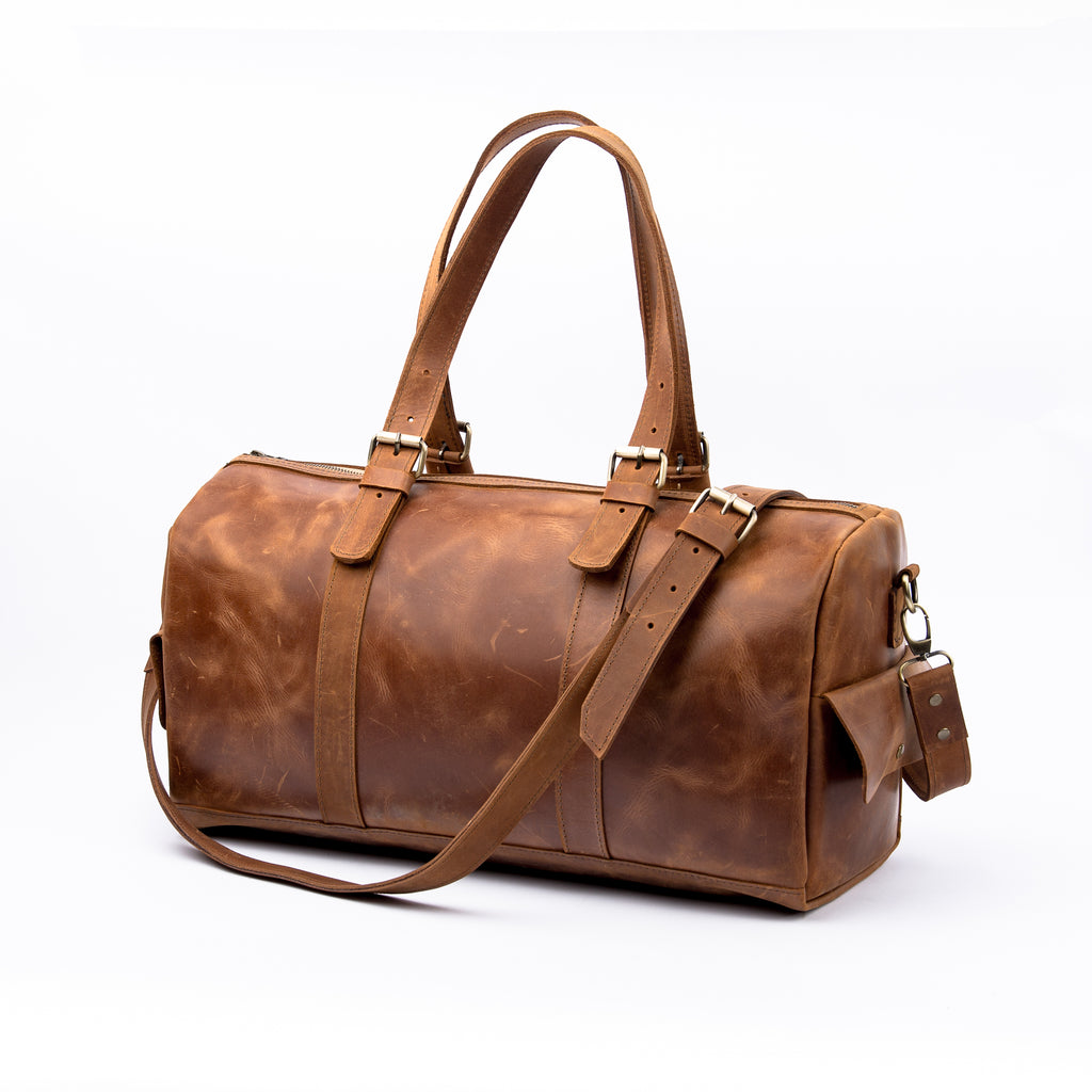 Front view of a vintage LIONELL brown leather duffle bag; handcrafted from premium genuine leather, featuring double top handles with brass buckle details and a matching detachable shoulder strap placed beside it, displayed on a clean white background.