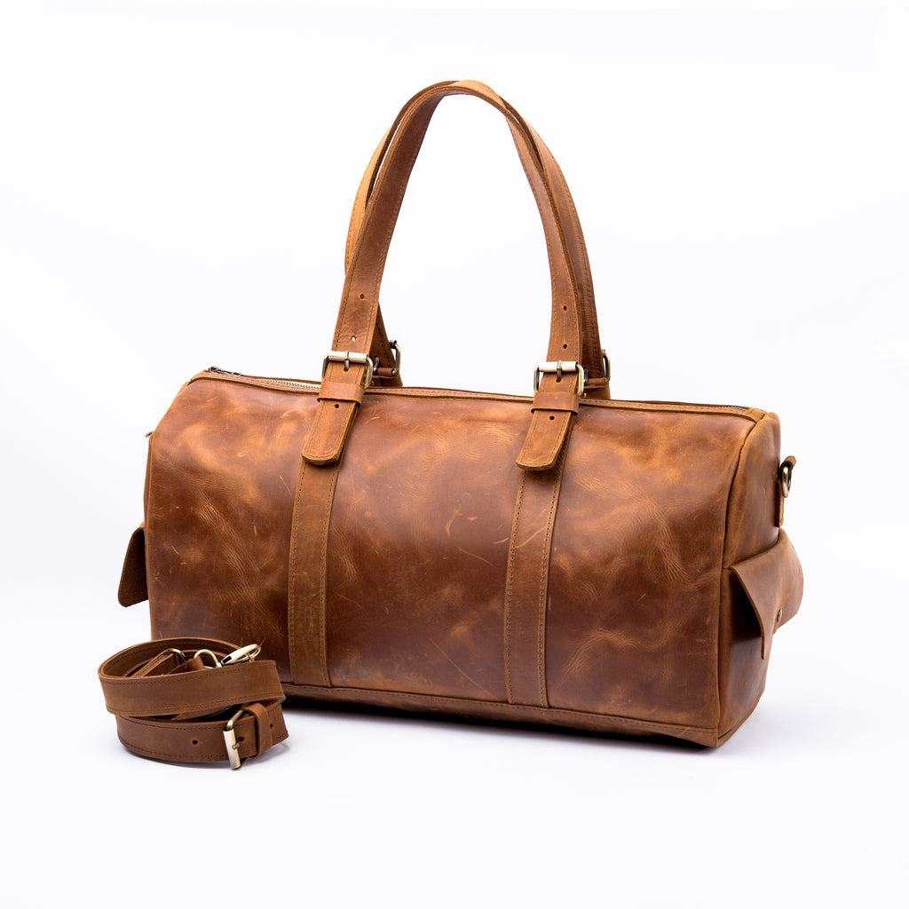 Side view of a LIONELL vintage brown leather duffle bag; handcrafted from genuine leather, featuring double top handles with brass buckle details and an attached leather shoulder strap, displayed on a clean white background.