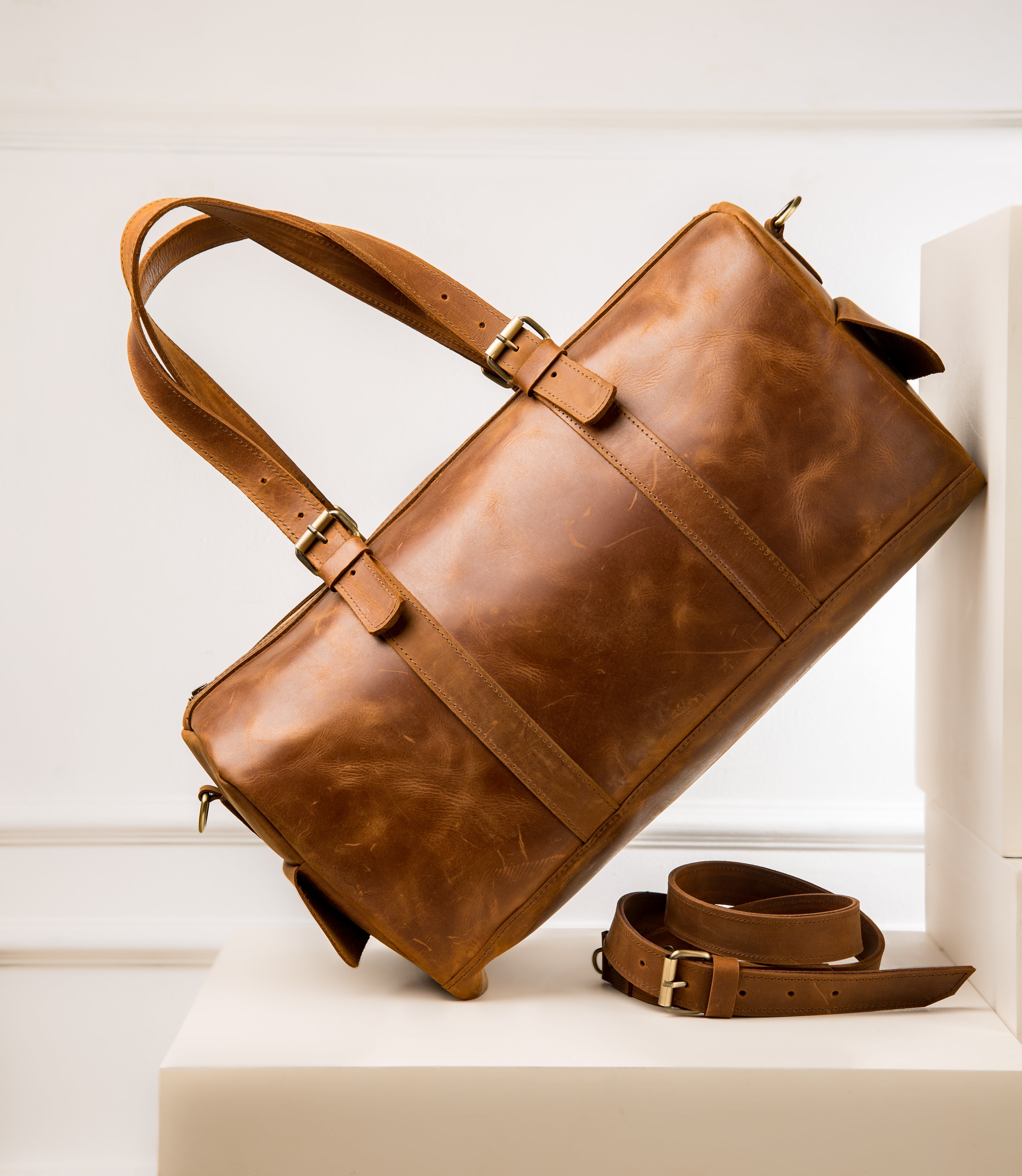 A photo of a vintage LIONELL brown leather duffle bag; handcrafted travel bag featuring double top handles with buckle details and a matching detachable shoulder strap, displayed diagonally on white blocks.